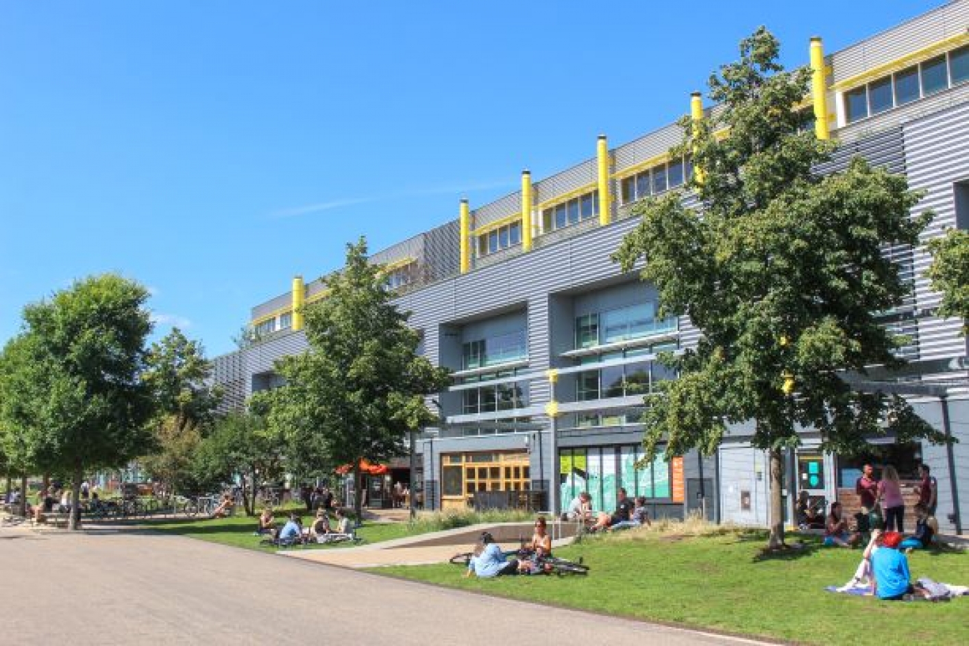 People sitting and reclining on te grass, surrounded by trees in front of building in a business park
